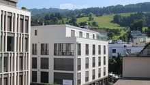 Modern multi-story building with large windows and balconies, surrounded by greenery and a scenic view of hills and trees.