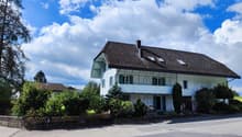 Two-story house with white exterior, dark roof, and balcony on the upper level. Surrounded by greenery and a driveway.