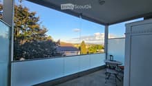 Balcony with glass railing, table, and chair, overlooking a scenic view of trees and a house.