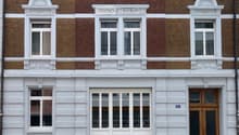 Classic European building facade with red brick upper levels and white plaster lower level, featuring multiple windows with decorative white surrounds and a central wooden door.