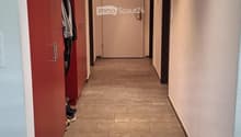 Clean hallway with tiled floor, beige walls, and a closed door at the end. A red wardrobe with hanging clothes is visible on the left side.