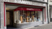 Facade of a shop named "Stampfli Optik" with a glass display window showing various optical items, located on a cobblestone street.