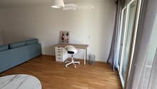 A modern, well-lit room with wooden flooring, a grey sofa, a white desk with a chair, a round marble table, and a hanging lamp.