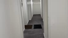 Empty hallway with white walls, gray carpeted floor, and recessed lighting. Two doors visible on the left side, with small rugs placed in front of them.