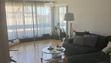 Modern living room with wooden flooring, large windows with sheer curtains, a dark grey sectional sofa with patterned cushions, a round glass coffee table, two floor lamps, and potted plants.