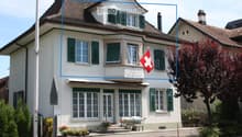 Two-story house with white walls, green shutters, and a red Swiss flag on the balcony. The house features a covered entrance with steps, potted plants, and a bench. The roof is tiled, and there are trees and shrubs in the surrounding garden area.