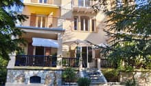 Two-story house with yellow and white exterior, multiple balconies, stone base, and lush greenery surrounding the property.