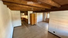 Empty room with wooden floor and exposed wooden ceiling beams. Adjacent kitchen area visible with cabinets and appliances.