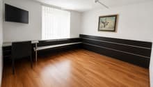 A modern, minimalist room with wooden flooring, a black chair, a small table, a mounted screen, and a framed picture on the wall.