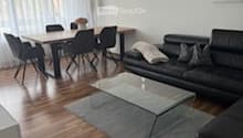 Bright, modern living and dining area with hardwood flooring, large windows with sheer curtains, a black sectional sofa, a glass coffee table, a dining table with six chairs, a floor lamp, and a decorative picture frame on the wall.