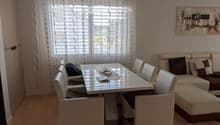 Bright living room with a sofa, coffee table, dining table, chairs, and a handbag on the floor. Large windows with white curtains and a modern painting on the wall.