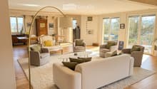 Modern living room with large windows offering a scenic view, featuring a cream-colored sofa, coffee table, armchairs, ottoman, floor lamp, dining table, and cabinet.