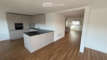 Modern kitchen with light grey cabinets, black countertop, and wooden flooring. Features a central island with a sink and faucet, and an adjacent living space with wooden flooring and a skylight.