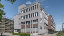 Modern multi-story building with large windows and brown accents, featuring signage for "Arbeitszentrum Liebfeld" and other companies. The building has a clean, contemporary design with a mix of light and dark tones.