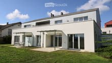 Modern two-story house with white exterior, large windows, and a balcony on the upper level. The house features a covered patio area with outdoor furniture and a well-maintained lawn.