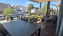 Balcony with a wooden table, chairs, a hanging wicker chair, potted plants, and an umbrella. The view shows a residential area with houses and trees.