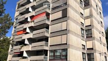 Modern multi-story apartment building with concrete facade, balconies, and large windows. Two cars are parked in front.