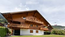 A large, multi-story wooden house with balconies and a spacious lawn in front, set in a scenic mountainous area.
