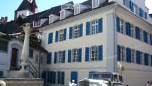 A large, multi-story building with white walls and blue shutters, featuring a classic car parked in front on a cobblestone street.