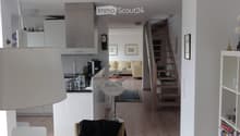 Open-plan living and dining area with a white dining table, chairs, a bar stool, a sofa in the background, a bookshelf, and a kitchen area with cabinets and appliances.