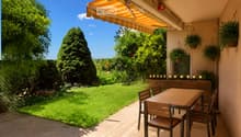 Balcony with a wooden table and chairs, surrounded by greenery and a well-maintained lawn.