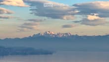 Panoramic view of a lake surrounded by mountains under a partly cloudy sky, with a few houses visible on the shore.