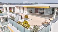 Modern rooftop terrace with glass railings, potted plants, and a view of the surrounding area.