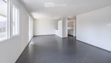 Empty room with white walls, dark tiled floor, large windows, and a small kitchen area with cabinets and appliances visible in the background.