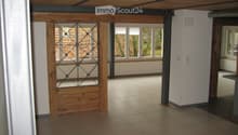 Empty room with tiled floor, white walls, wooden ceiling beams, and large windows with metal-framed glass panels.