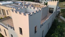 Modern two-story building with a distinctive, castle-like design featuring crenellated roofline and multiple chimneys. The structure includes a wooden deck area, glass railing, and surrounding greenery.