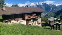 A wooden chalet with a brown tiled roof, wooden balcony, and stone base, situated on a grassy slope with a stunning mountain backdrop.