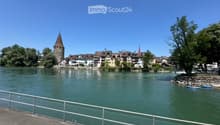 Scenic riverside view with historic buildings, a tower, and a small boat on calm water.