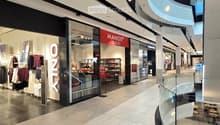 Shopping mall interior with glass storefronts, clothing racks, shelves, and a cashier counter visible inside a store.