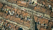 Aerial view of a historic European city with numerous buildings featuring red-tiled roofs, narrow streets, and a prominent church spire in the center.