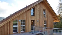 A modern wooden house with a sloped roof, large windows, and a raised ground floor accessed by a staircase.