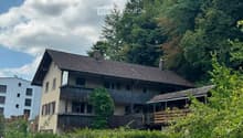 Two-story house with a tiled roof, multiple windows, and wooden balconies surrounded by lush greenery and trees.
