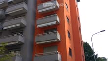 Modern multi-story apartment building with orange and grey facade, multiple balconies, and a gated entrance.
