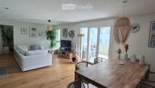 Bright living and dining area with wooden flooring, white sofa, dining table with chairs, cat furniture, and large windows leading to a balcony.
