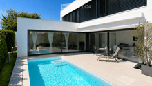 Modern two-story house with large glass sliding doors, a swimming pool, outdoor seating area with lounge chairs and a small table, and a well-maintained garden.