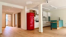 Open-plan kitchen and living area with light wooden flooring, white cabinets, a red refrigerator, a wooden table, and built-in shelving.
