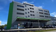 Modern multi-story building with grey facade and green accents, featuring a large truck parked in front and several cars in the parking area.