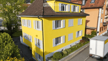 Bright yellow two-story house with white shutters and multiple windows, surrounded by greenery and a neighboring building.