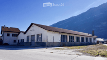 A two-story building with a flat roof and large windows, situated in a rural area with a mountain in the background.