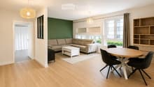 Modern living and dining area with light wood flooring, a beige sectional sofa, a white coffee table, a round wooden dining table with four black chairs, and built-in wooden shelving units.