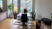 Dining area with a wooden dining table, six chairs, a sofa, a cabinet with electronic devices, and a hanging lamp. Large windows and sliding doors lead to a balcony with plants.