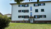 Multi-unit apartment building with white exterior, green shutters, and a garden in the front.