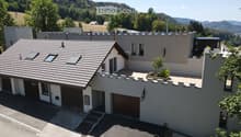 Modern two-story house with a tiled roof, garage, and a small balcony area with plants. The house is surrounded by a scenic, green landscape with hills in the background.