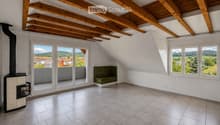Bright, empty attic room with wooden beams, large windows, and a wood-burning stove. The space features white paneled walls, tiled flooring, and a small seating area with a green cushioned bench.