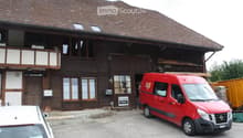 Traditional wooden building with a steeply pitched roof, large windows, and a balcony. A red van is parked in front, and there is a sign on the building.