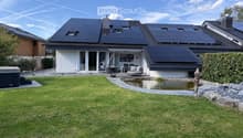 A modern house with solar panels on the roof, surrounded by a well-maintained lawn, a small pond, a patio area with outdoor furniture, and a hot tub.